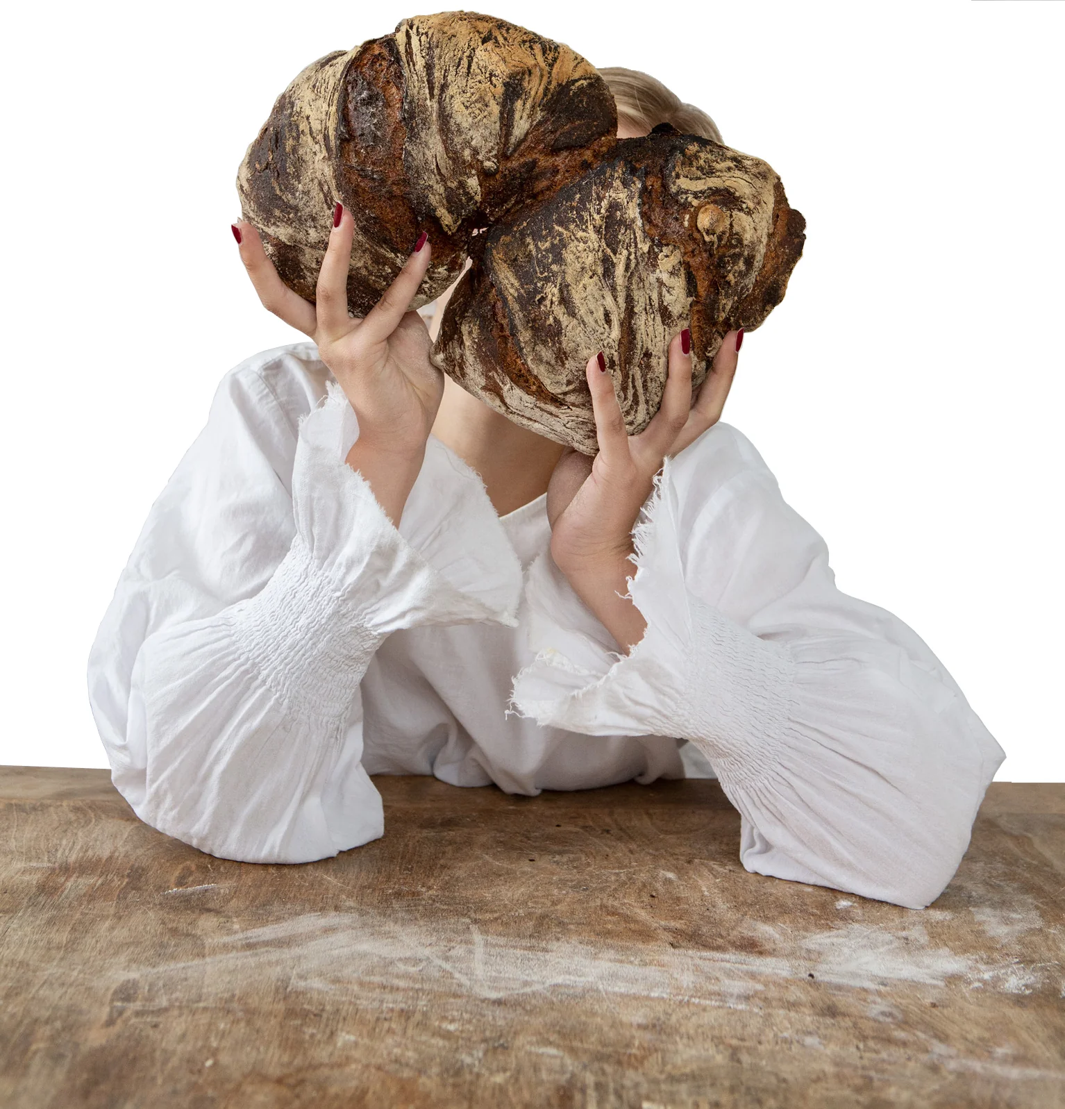 A woman in a white blouse holds two large round sourdough loaves up in front of her face while leaning on a wooden table.